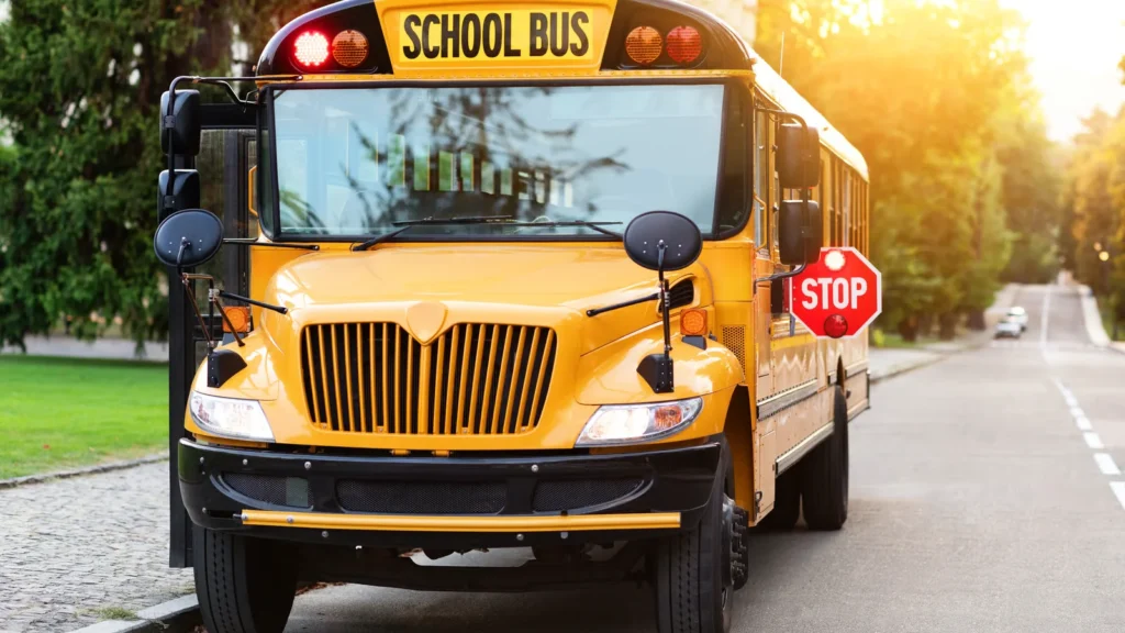 A realistic image of a yellow school bus stopped on a U.S. road with flashing red lights and a stop arm extended while cars wait behind it.